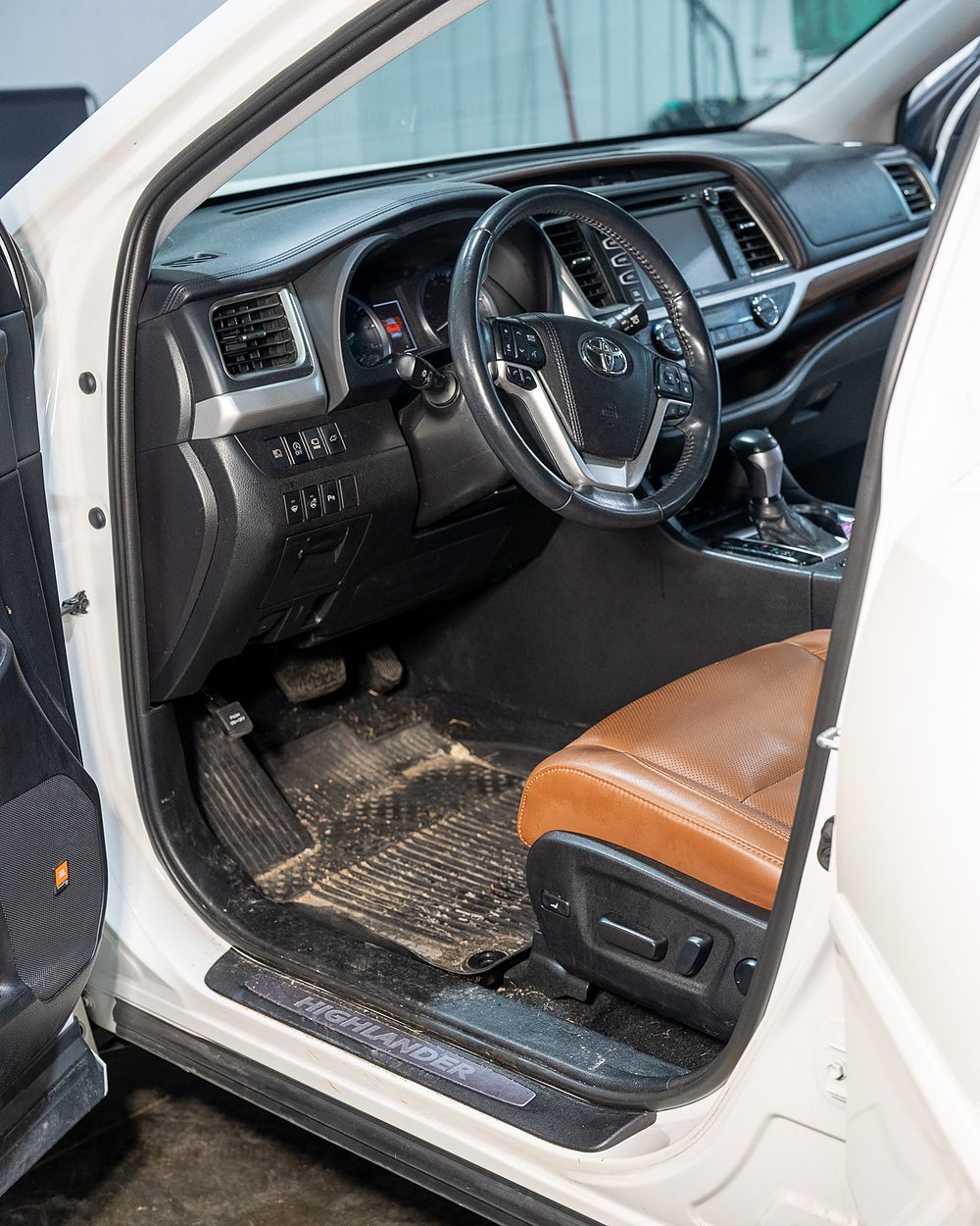 Front interior of a white Toyota Highlander before detailing, with a dirt-filled floor mat, dust through the dash and console, and grime along the door sill