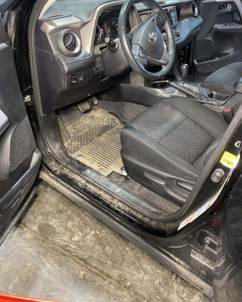Driver footwell and door sill of a black Toyota RAV4 before detailing, caked with white road salt and grit across the mat, sill, and rocker panel