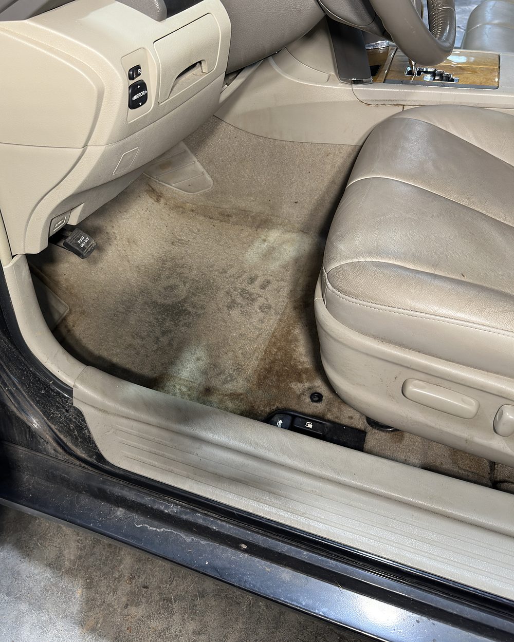 Driver footwell of a sedan before detailing, the light beige carpet stained grey and brown across the whole floor and up onto the door sill