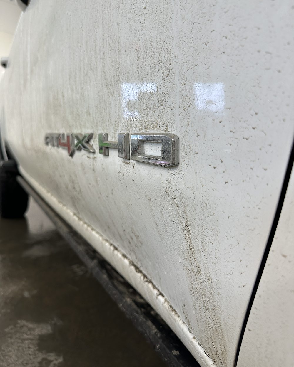 Door and rocker panel of a white pickup before washing, under a film of road grime and salt spray with dirt packed around the chrome badge